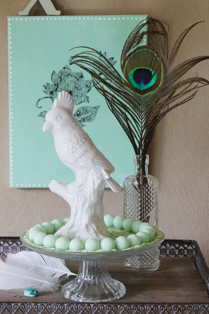 White chain cockatoo and string of turquoise beads on glass cake stand in front of peacock feather in vase