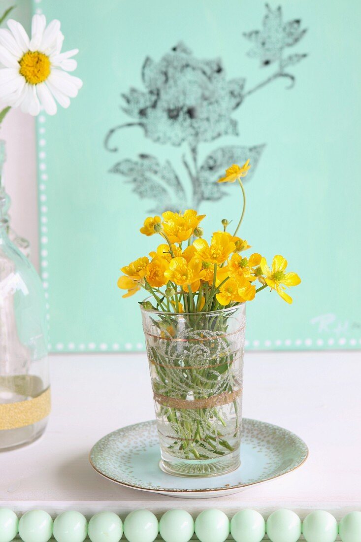 Gelbe Wiesenblumen im Glas auf Untertasse vor grüner Leinwand mit bedrucktem Blumenmuster, im Vordergrund grüne Perlenkugeln