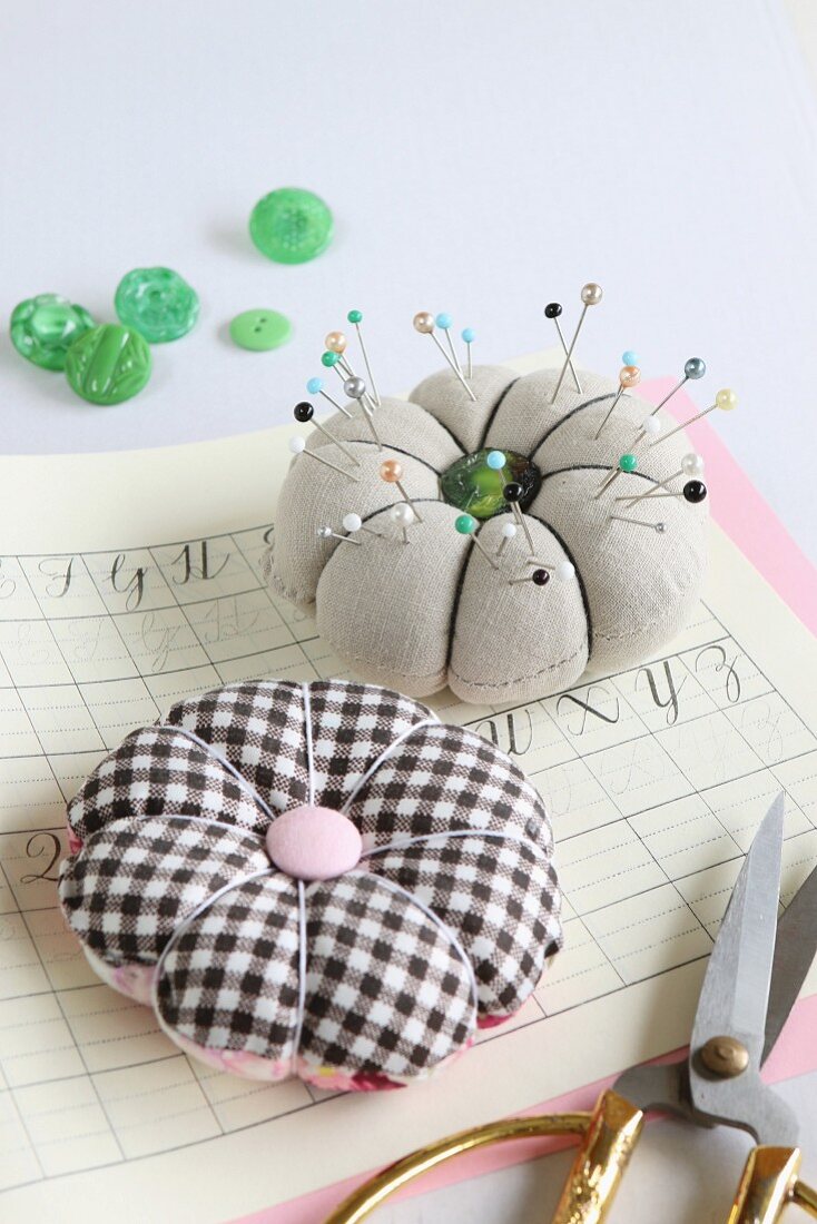 Pins in hand-made, flower-shaped pin cushions on exercise book and various green buttons in background