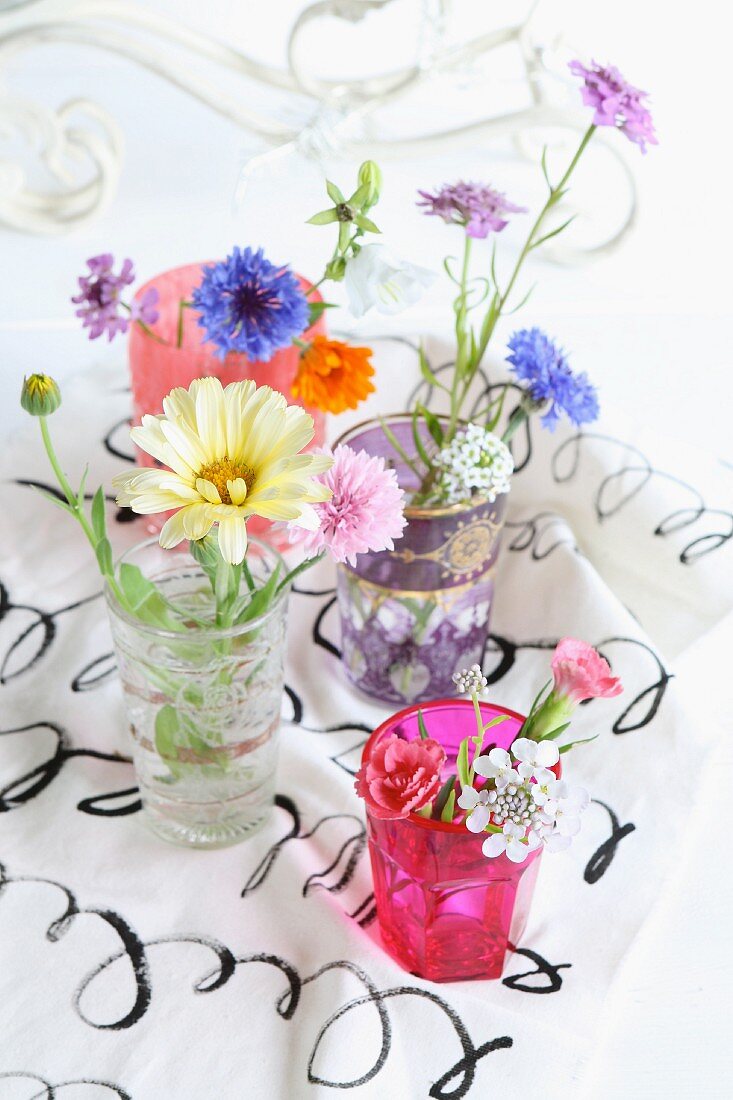Stillleben mit bunten Wiesenblumen in unterschiedlichen Gläsern auf bedrucktem Stoff
