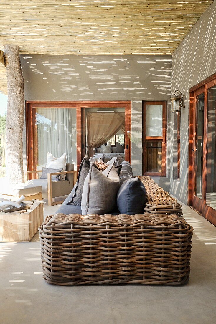 Sofa with chunky wicker frame on veranda with grey-painted walls and stripped wooden ceiling