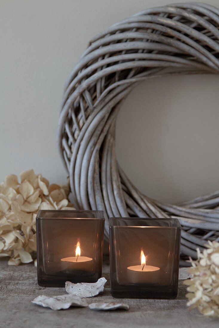 Lit tealights in smoked glass tealight holders in front of wicker wreath painted pale grey