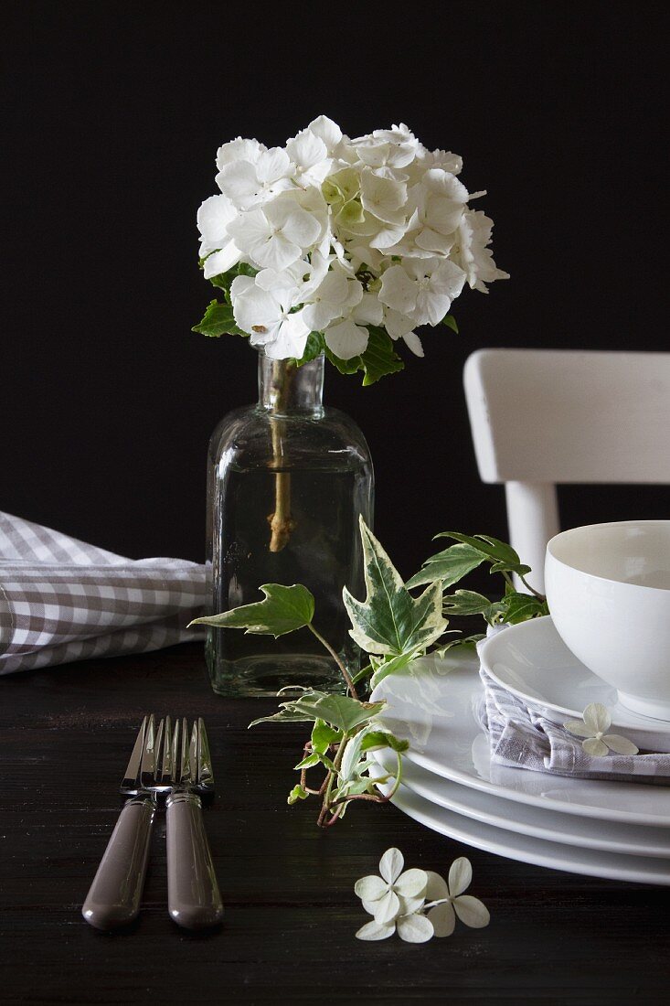 White crockery, sprig of ivy and white hydrangea in vintage glass bottle against dark background