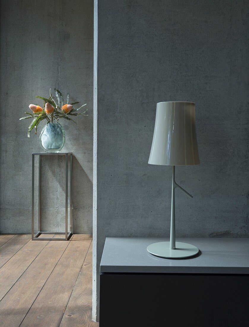 Table lamp on bedside cabinet against exposed concrete wall and vase of exotic flowers on small metal table in background