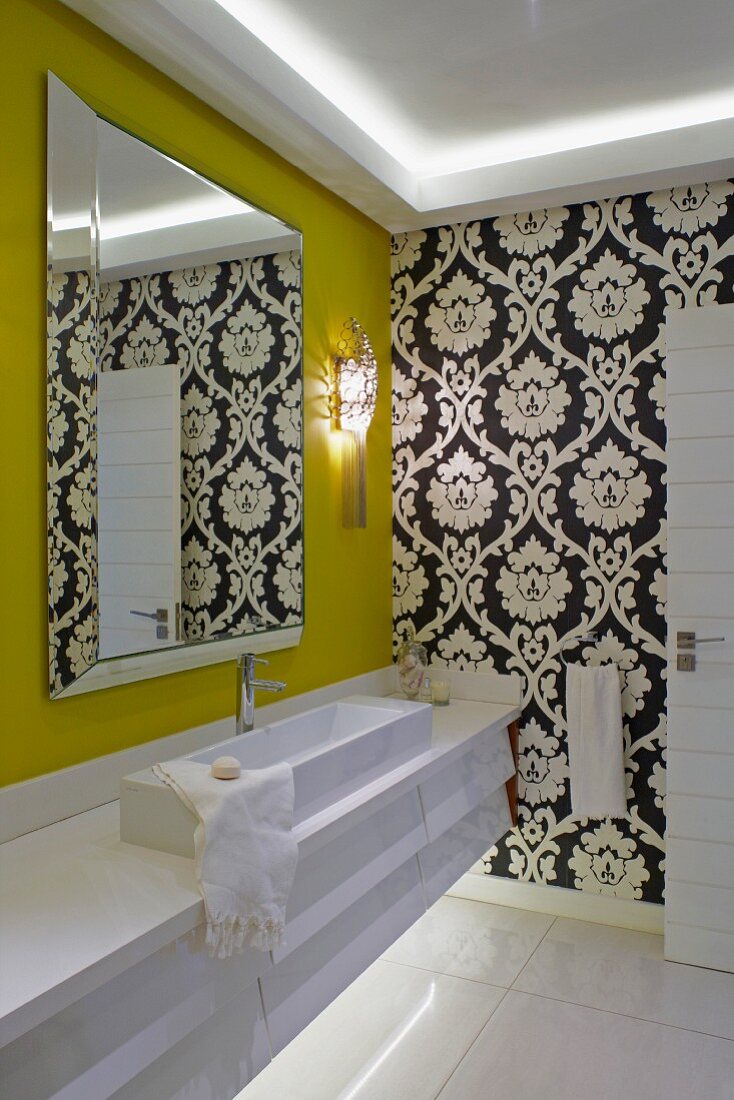 Moderner Waschtisch mit länglichem Aufsatzbecken und geschliffenem Spiegel auf senfgelber Wand, im Hintergrund klassische schwarz-weiße Tapete