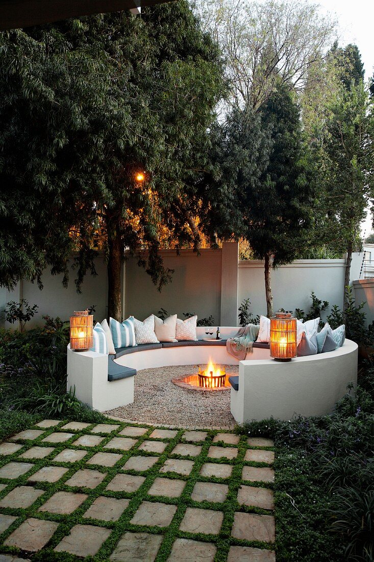 Gemütlicher, kreisförmiger Sitzplatz mit Kissen um Feuerstelle im Garten, Laternen auf gemauerter Rückenlehne in Abendstimmung
