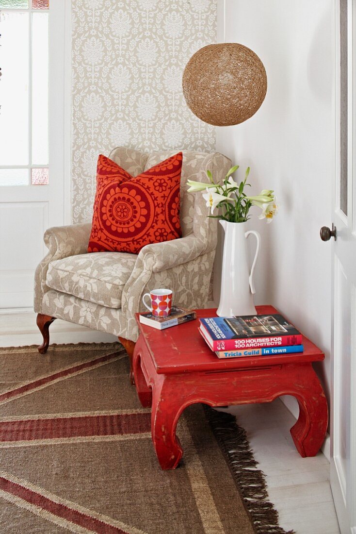 Reading corner; opium table and pale armchair with patterned scatter cushions against wallpapered wall