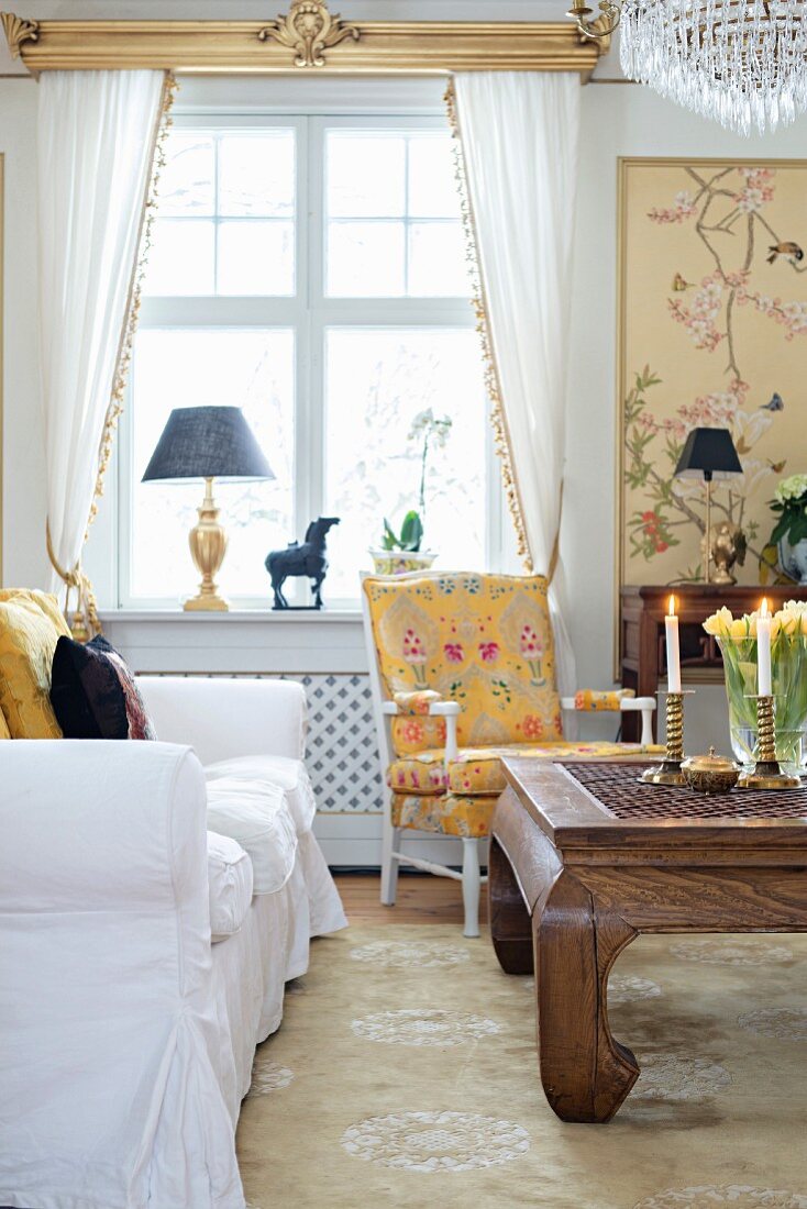 Wohnzimmer mit weißem Sofa, antikem Couchtisch aus Massivholz, im Hintergrund Armlehnsessel vor Fenster mit weissen, drapierten Vorhängen
