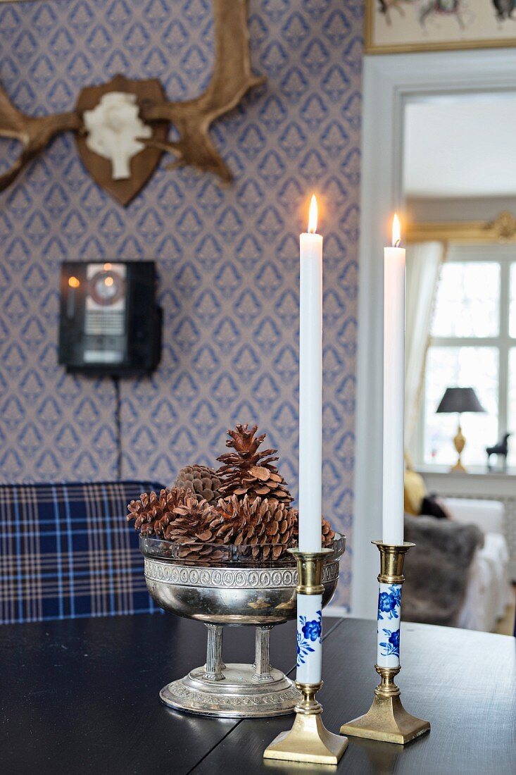Kerzenhalter und Tannenzapfen in antiker Silberschale auf Tisch, im Hintergrund Hirschgeweih an blauer Tapete