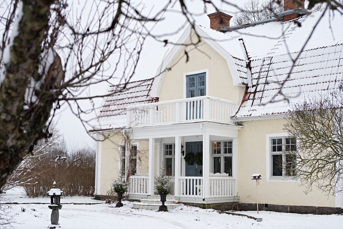 Blick vom winterlichen Garten auf renoviertes Landhaus mit weißem Holzgeländer an Veranda und auf Balkon