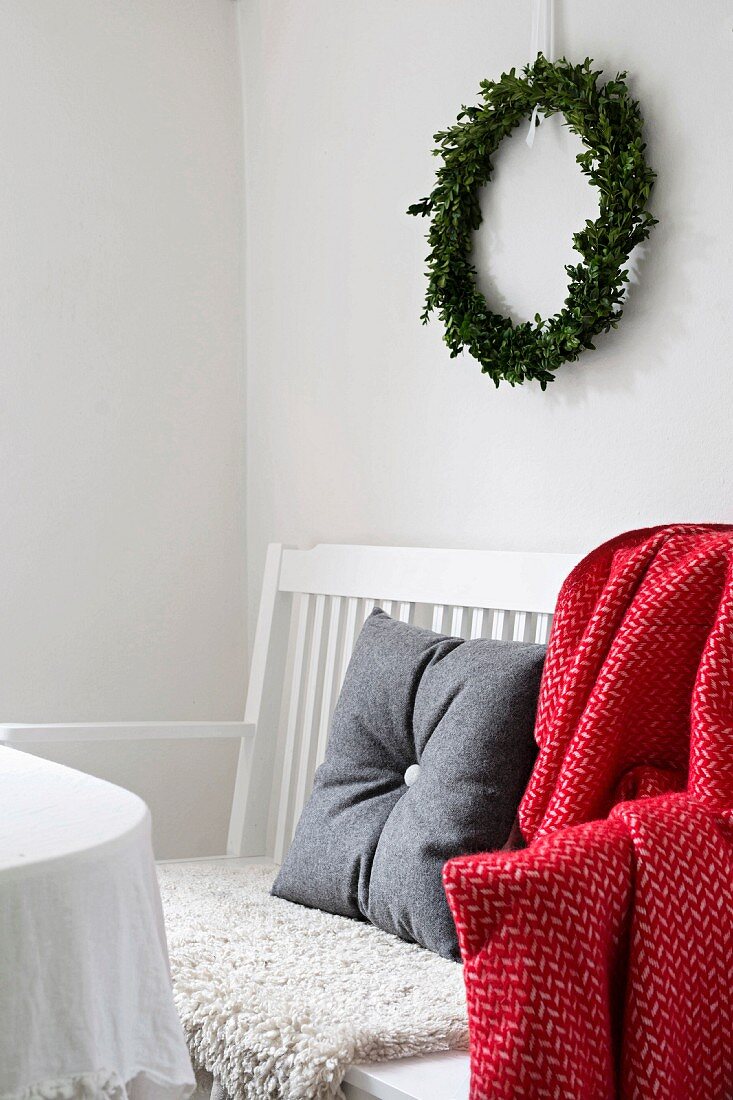 Grau bezogenes Filzkissen und rot weiss gemusterte Decke auf weiss lackierter Sitzbank, unter weihnachtlichem Wandkranz
