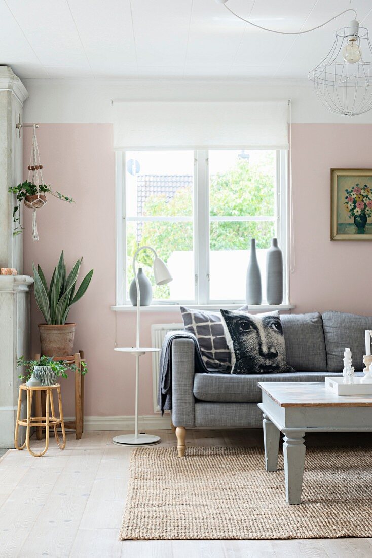 Kissen mit Gesichtsmotiv auf Polstersofa vor Fenster, seitlich Blumentöpfe auf schlichten Hockern in rosa getöntem Wohnzimmer mit romantischem Flair