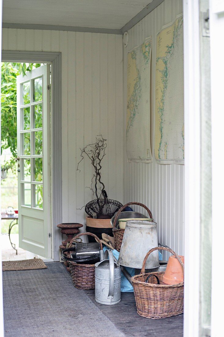 Weidenkörbe und Gartenutensilien auf Boden im Hausflur, weiße Holzverschalung an Wand, im Hintergrund offene Gartentür