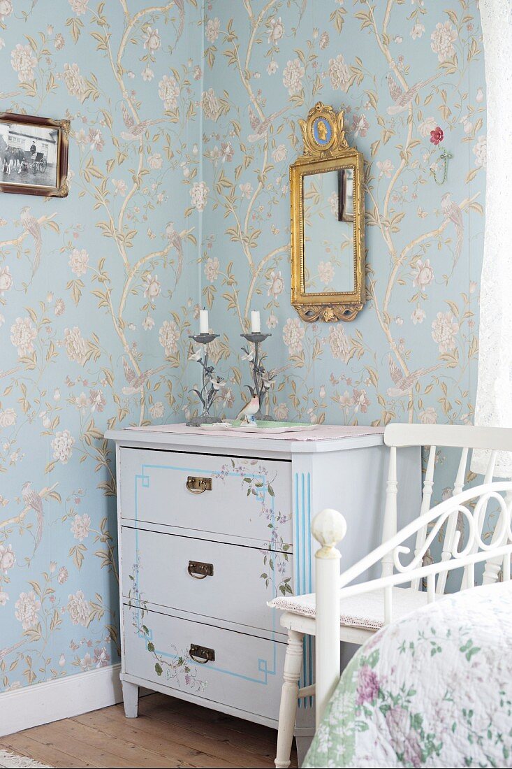Bemalte Kommode in Zimmerecke, an Wand floral gemusterte Tapete und Spiegel mit verziertem Goldrahmen