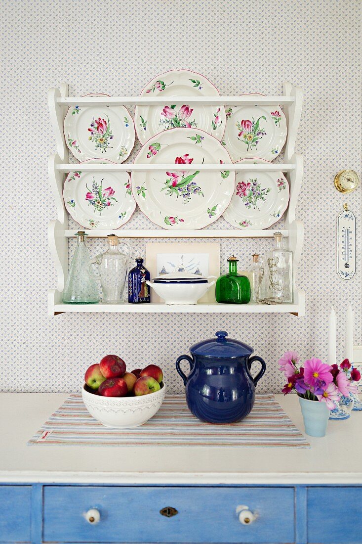 Detail of fruit bowl and blue ceramic pot on kitchen counter below decorative wall plates in plate rack mounted on wallpapered wall