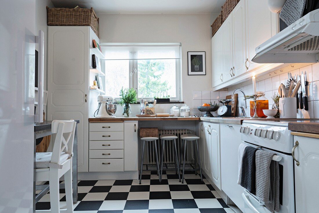 Bar stools at dining area below window and black and white chequered floor in white, Scandinavian fitted kitchen