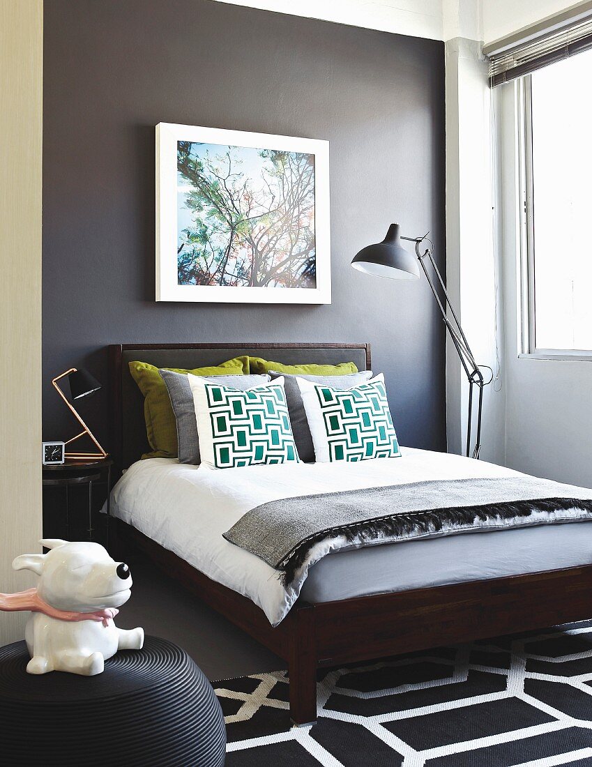 Bedroom with wall painted dark grey, scatter cushions on double bed and dog figurine on pouffe in foreground