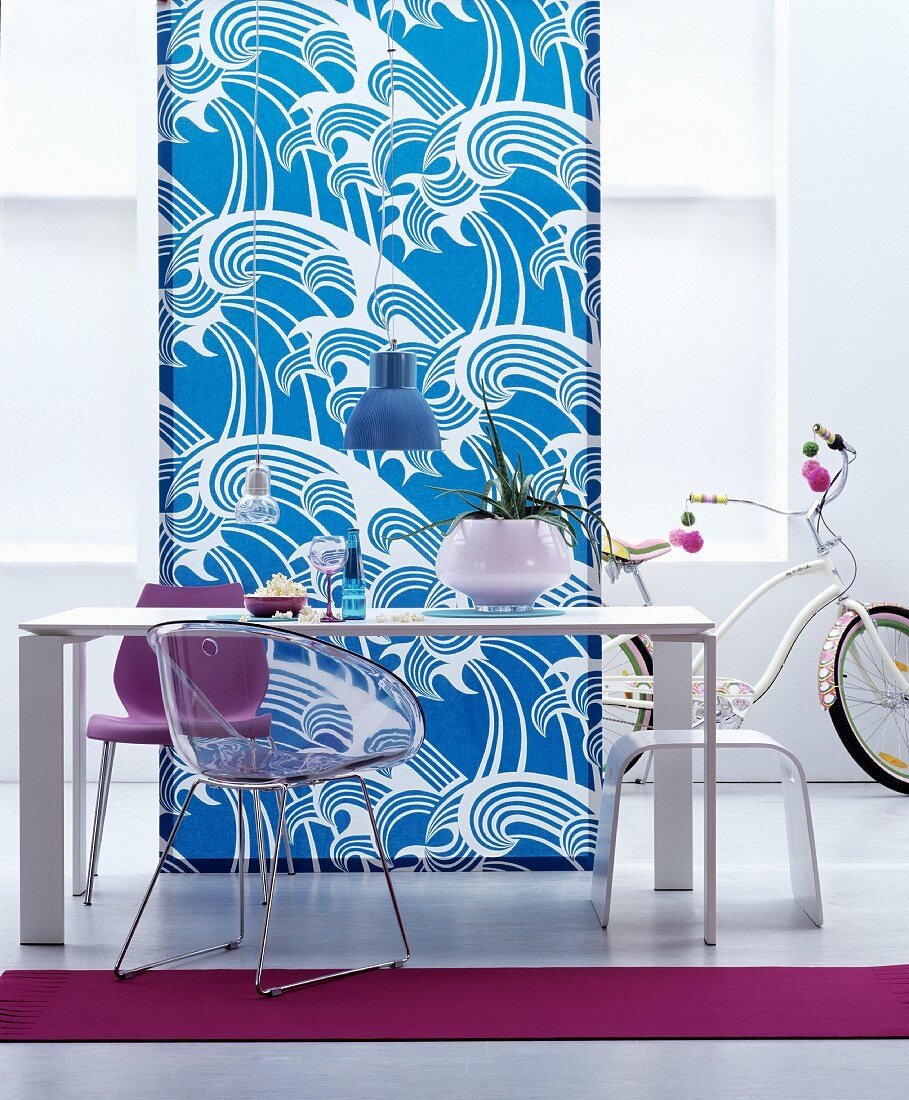Simple white dining table, stool and chairs in front of length of fabric with pattern of stylised waves