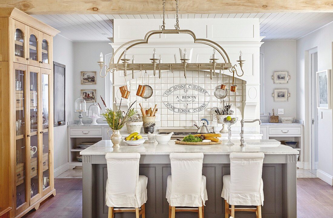 Bar stools with loose covers at island counter below kitchen utensils hung from pendant lamp in country-house kitchen