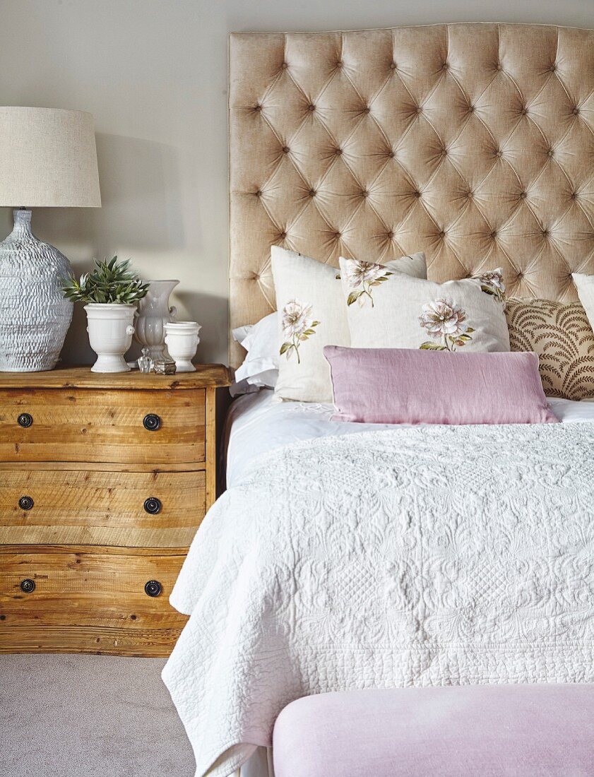 Double bed with button-tufted headboard, structured bedspread and floral scatter cushions next to rustic, county-house chest of drawers
