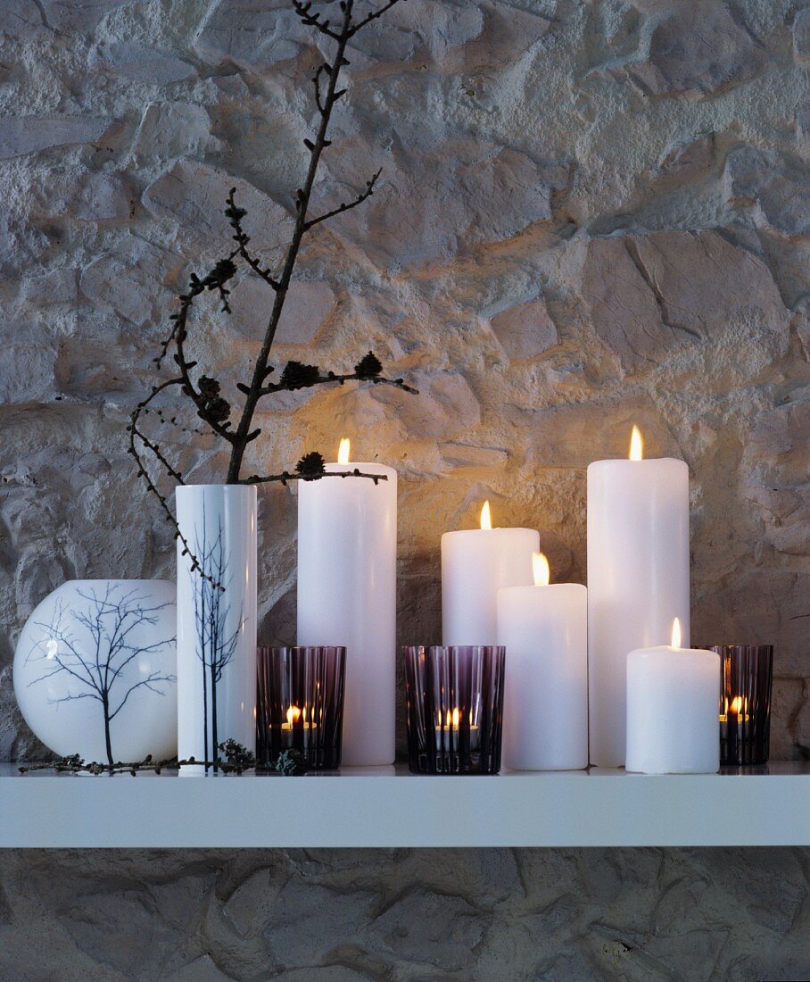 Stimmungsvolle winterliche Dekoration aus Vasen, Windlichtern und Stumpenkerzen auf weißem Wandregal