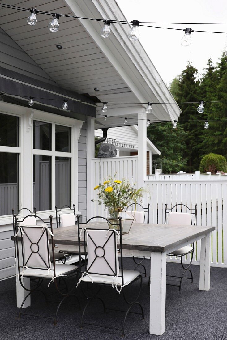 Elegante Metallstühle mit weissen Polstern um schlichten Holztisch, auf Terrasse mit grauem Bodenbelag vor Holzhaus