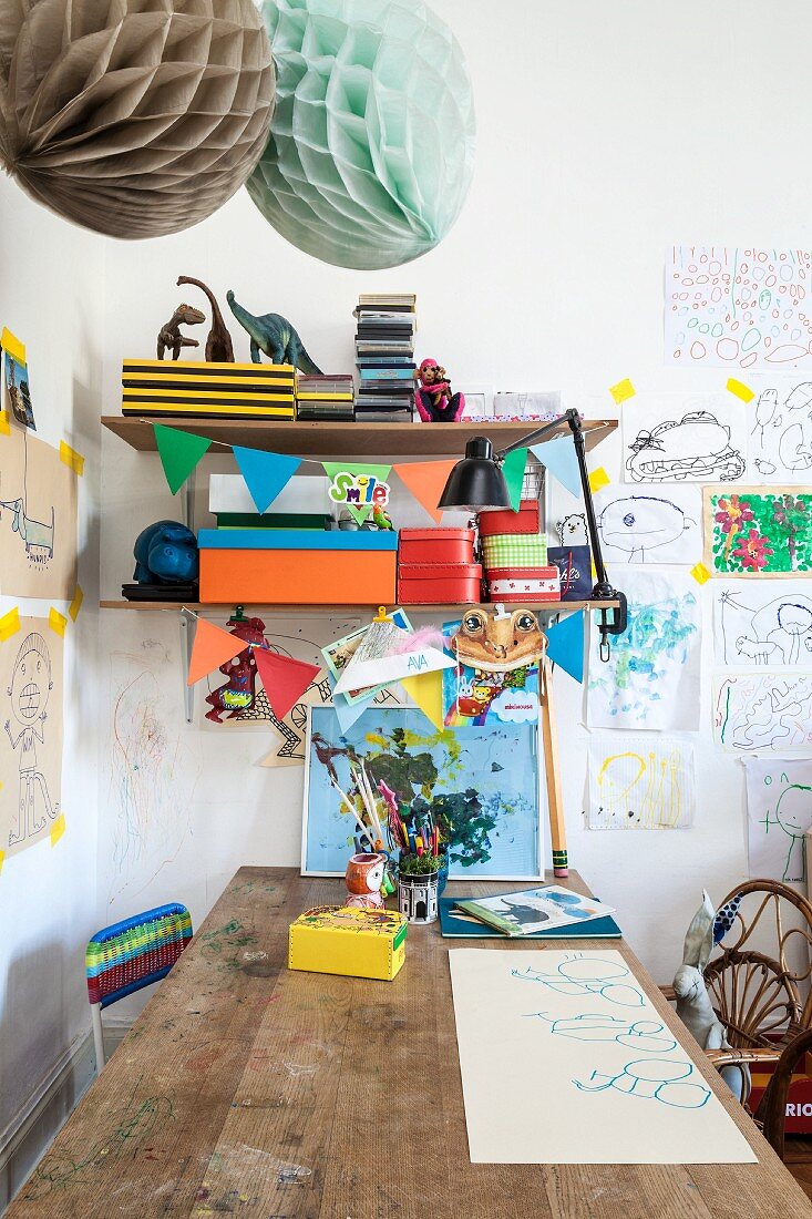 Holztisch vor Wand, oberhalb Ablagen mit farbigen Schachteln zwischen Kinderzeichnungen an Wand