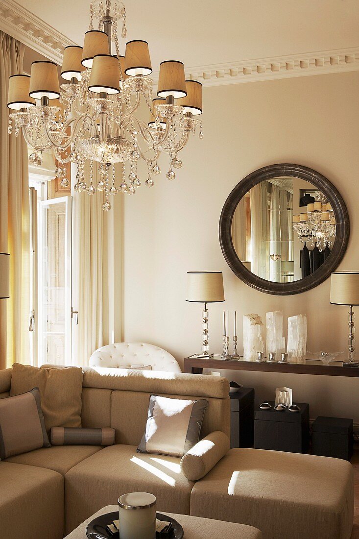 Crystal chandelier above sofa combination, elegant ornaments and classic table lamps on console table below round mirror on wall