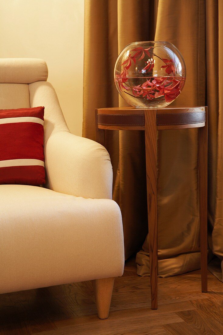 Branch of red leaves in spherical vase on elegant side table, floor-length curtains and armchair with red and white scatter cushion