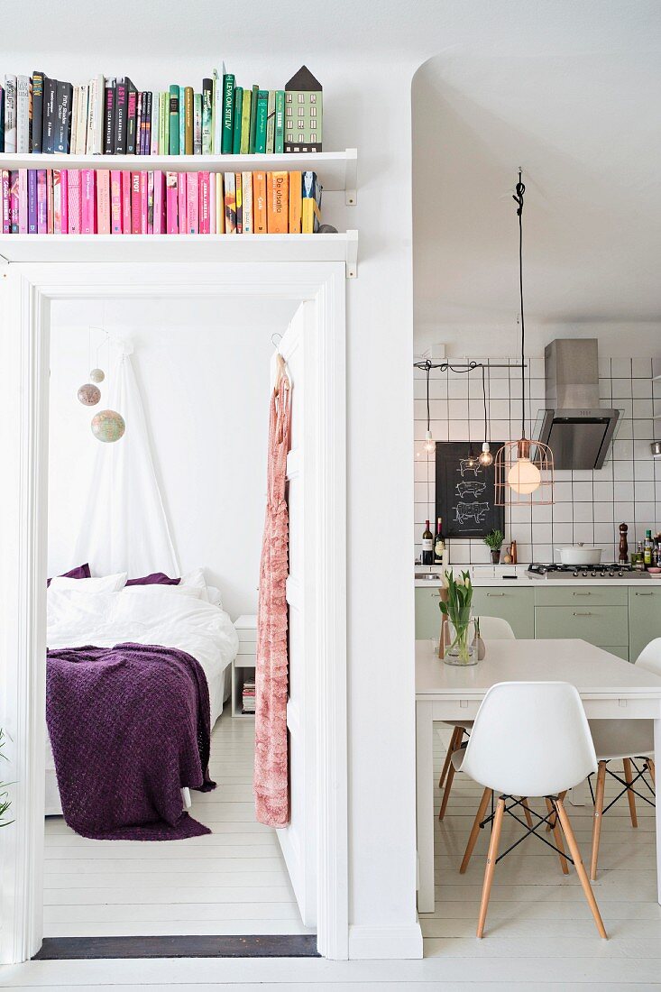 Bücherregal über offener Schlafzimmertür und Blick auf Bett mit violetter Tagesdecke, seitlich offene Küche mit Essplatz
