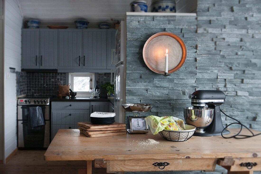 Blick über Esstisch aus Holz auf Naursteinwand, mit Wandkerzenhalter, seitlich Nische mit eingebauter Küche