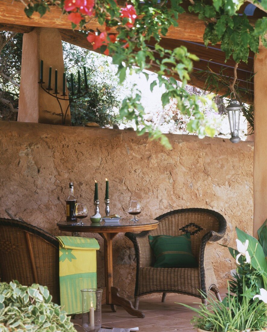 Romantischer Sitzplatz, Kerzenlicht und Wein auf Tisch, gemütliche Rattansessel vor Natursteinwand auf Terrasse