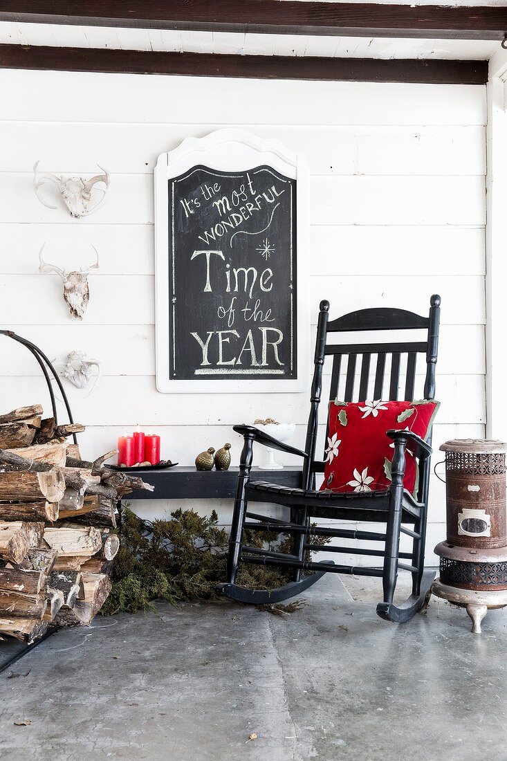 Red floral cushion on rocking chair, stacked firewood and candles on roofed veranda: motto on chalkboard on wall