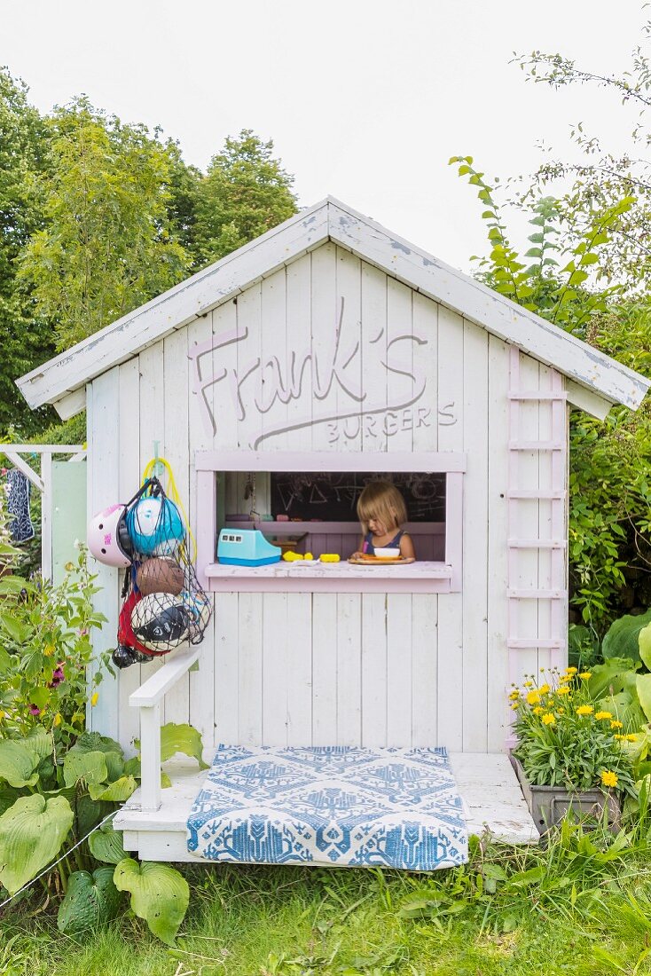 Giebelfassade eines Holz-Spielhauses in sommerlichem Garten und spielendem Mädchen