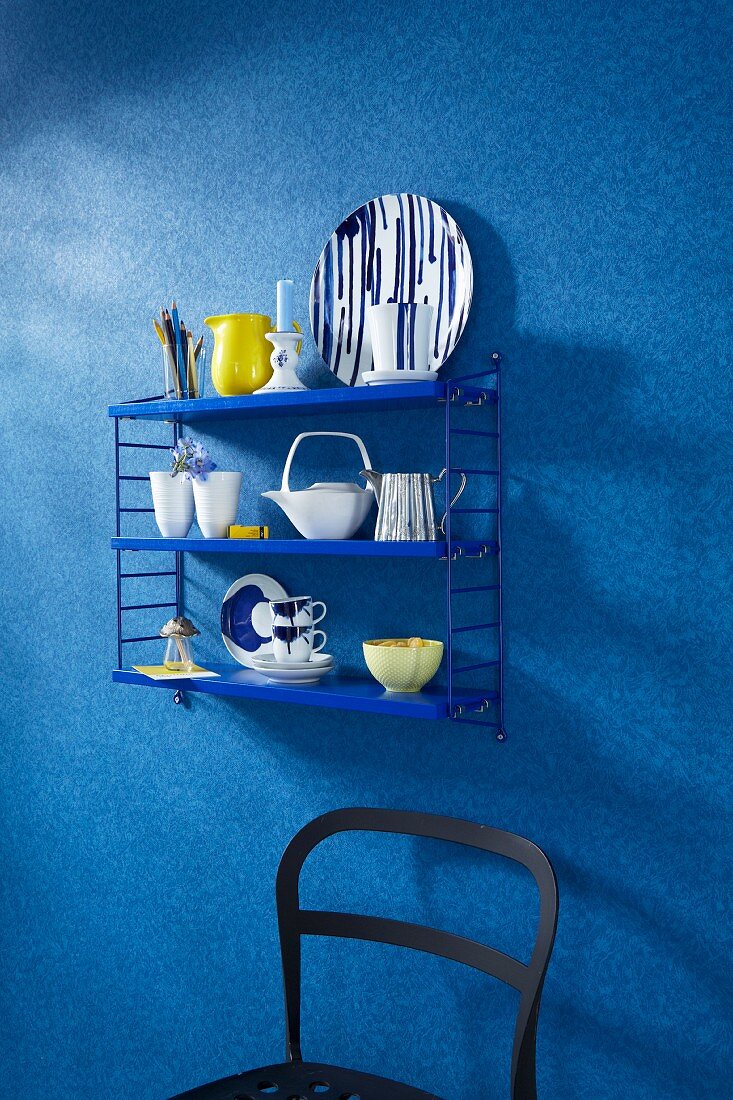 Blaues Stringregal mit Geschirr über teilweise sichtbaren schwarzen Stuhl, an tapezierter Wand mit Faux-Unis Muster in Blau