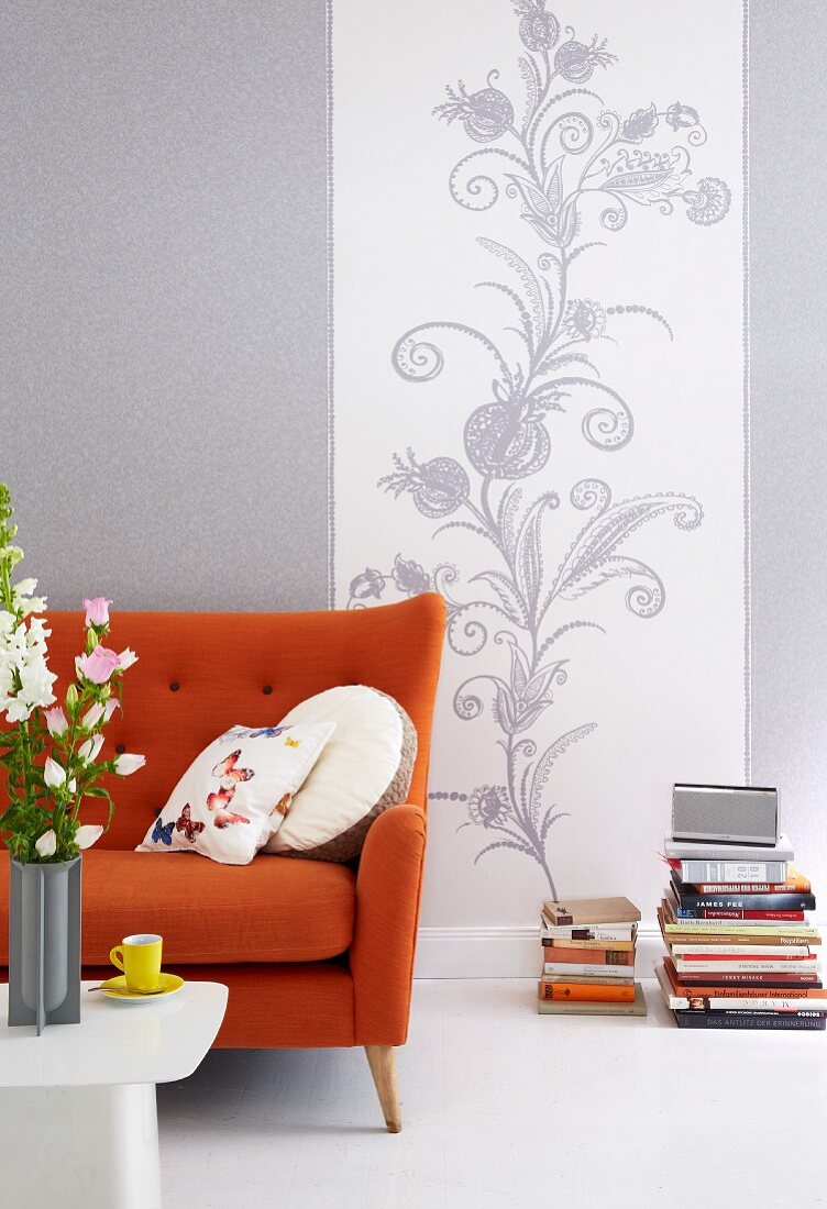 An orange-coloured sofa against a wall papered with a grey and white vine pattern and a grey semi-solid pattern