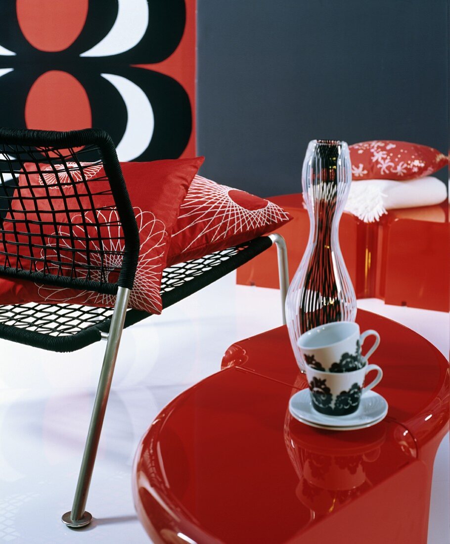 Retro-style arrangement of black chair with cord seat, cups on red side table and red, white and black picture on wall
