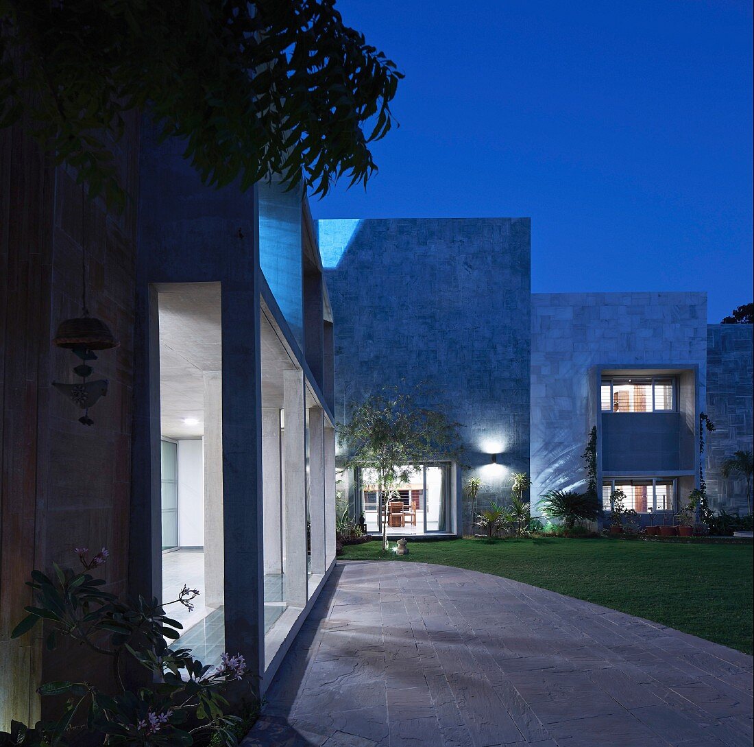 Tall, illuminated stone façade with terrace doors, garden, stone-paved area in front of concrete arcade at twilight