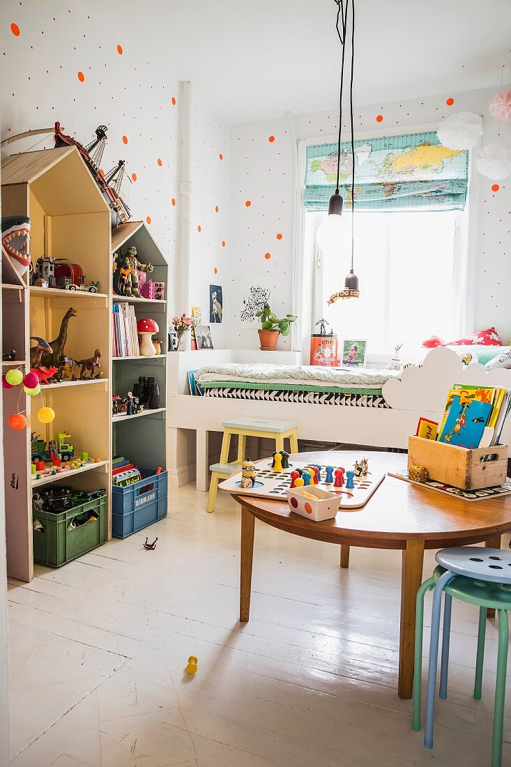 Runder Holztisch mit Spielsachen auf weißem Dielenboden, seitlich Regale in Hausform, neben weißem Bett am Fenster im Kinderzimmer
