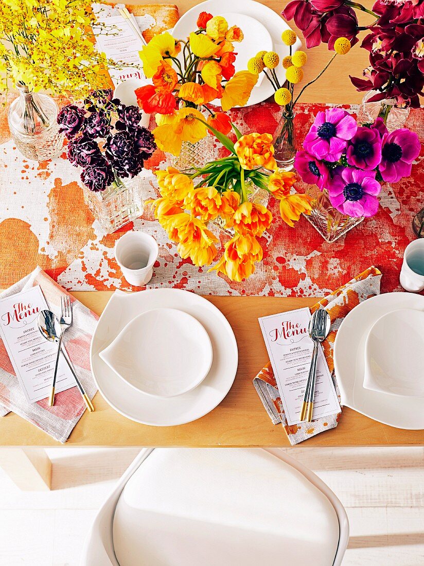 Gedeckter Tisch mit üppiger Blumendeko in Gelb- und Rottönen, Menükarten und gesprenkeltem Tischläufer