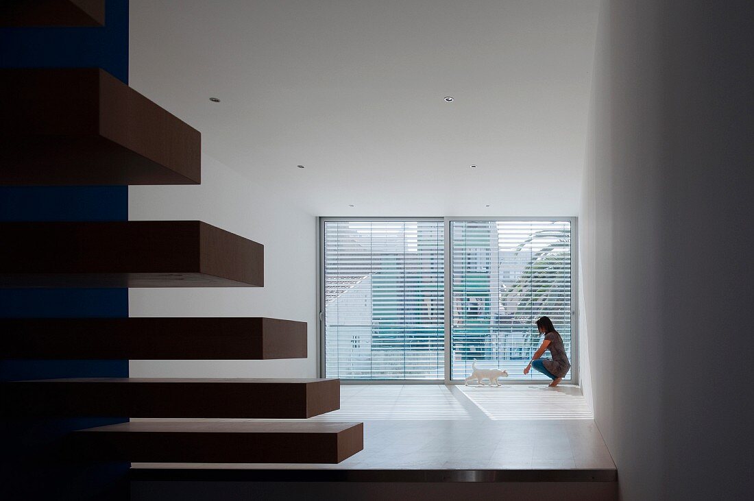 View past cantilever stair treads to woman in background next to large windows with open blinds