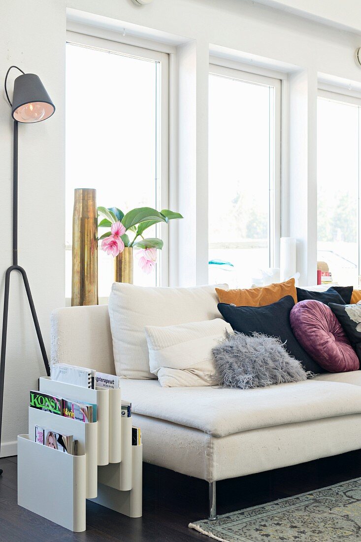 Zeitschriftenständer und Designer-Stehlampe neben heller Couch mit Kissenlager vor Fenster