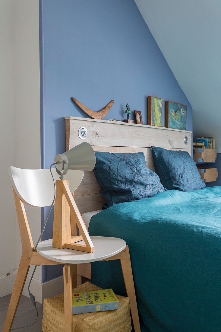Double bed with rustic wooden headboard against blue wall, blue satin bed linen and table lamp on designer wooden chair