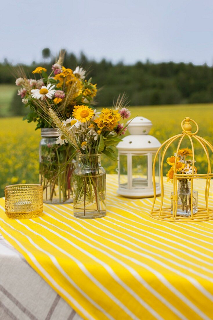 Bunte Wiesenblumensträusse in Vintage-Gläsern, Dekokäfig und Laterne auf gelbweiss gestreiftem Tischtuch in Freiem