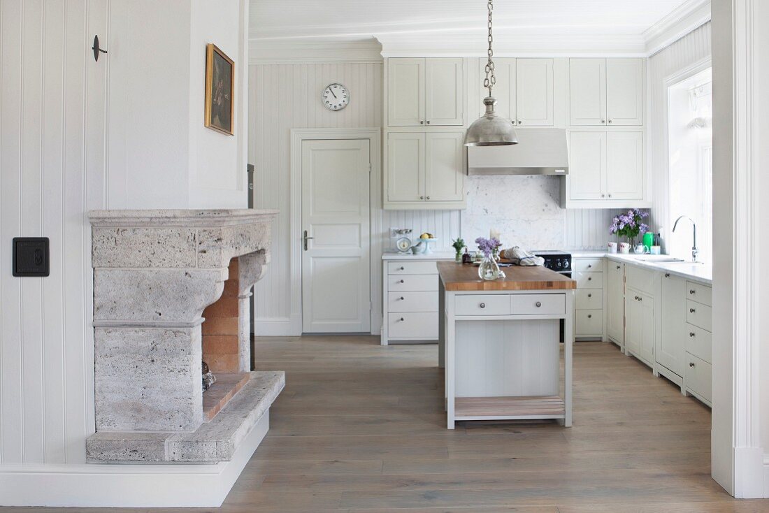 Spacious, white, country-house kitchen with free-standing island counter below metal pendant lamp and open fireplace with stone surround