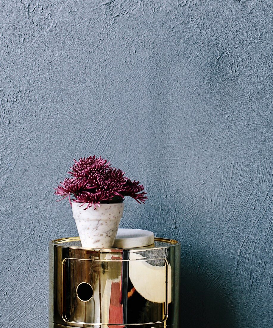 Dark red flowers in a marble plant pot on a shiny gold container against a gray wall