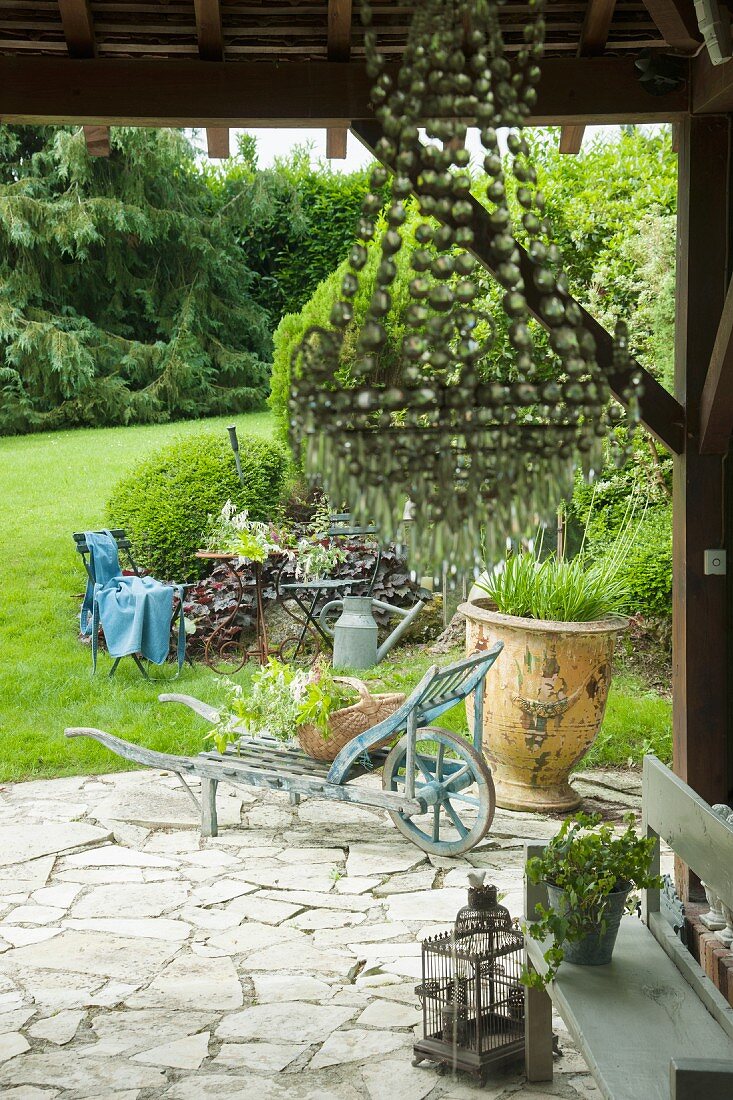 Vintage Schubkarre und Pflanzgefäss auf Terrasse mit Natursteinbelag, im Vordergrund Pendelleuchte mit Glasschmuck an Holzdecke