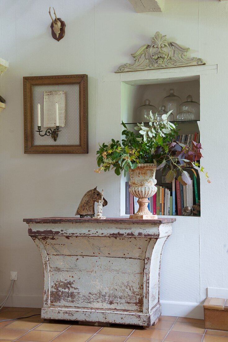 Leafy branches in antique planter on top of vintage console table with peeling paint between niche in wall and candle sconce in empty picture frame