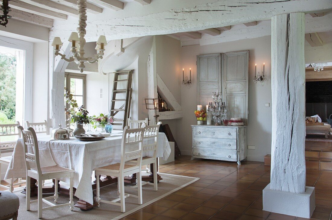 White chairs with carved backrests, table with white tablecloth and whitewashed supporting structure in dining area of old country house next to window