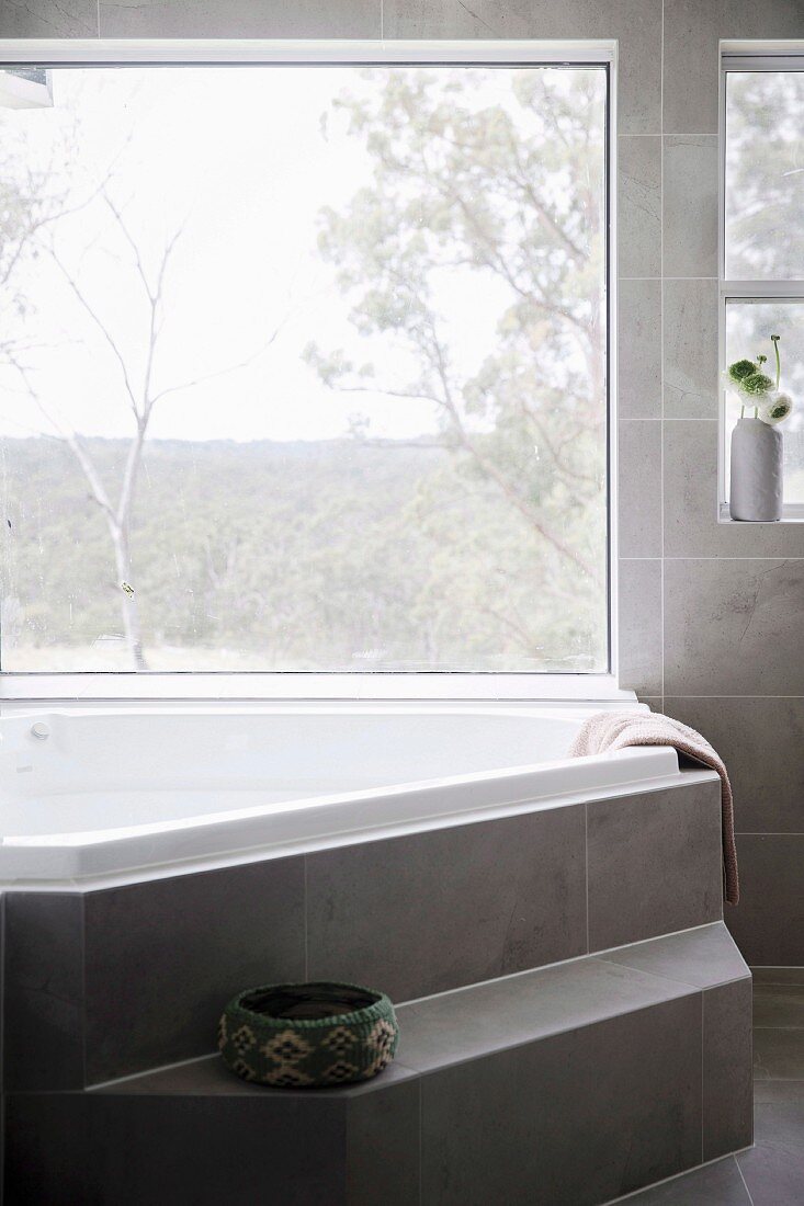 Polygonale Badewanne mit Stufe vor Panoramafenster, an Wand und auf Boden grau marmorierte Fliesen